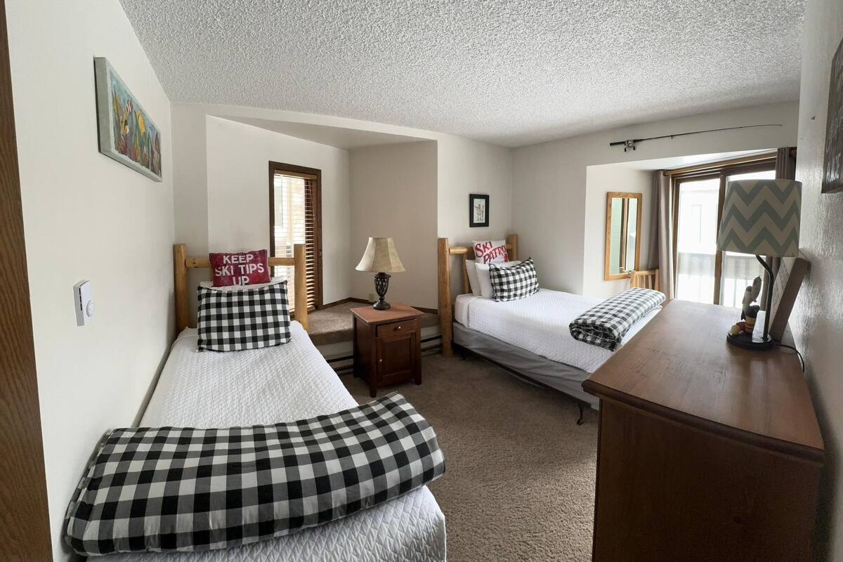 A room with two beds, a wooden dresser, a lamp, and a window with curtains.