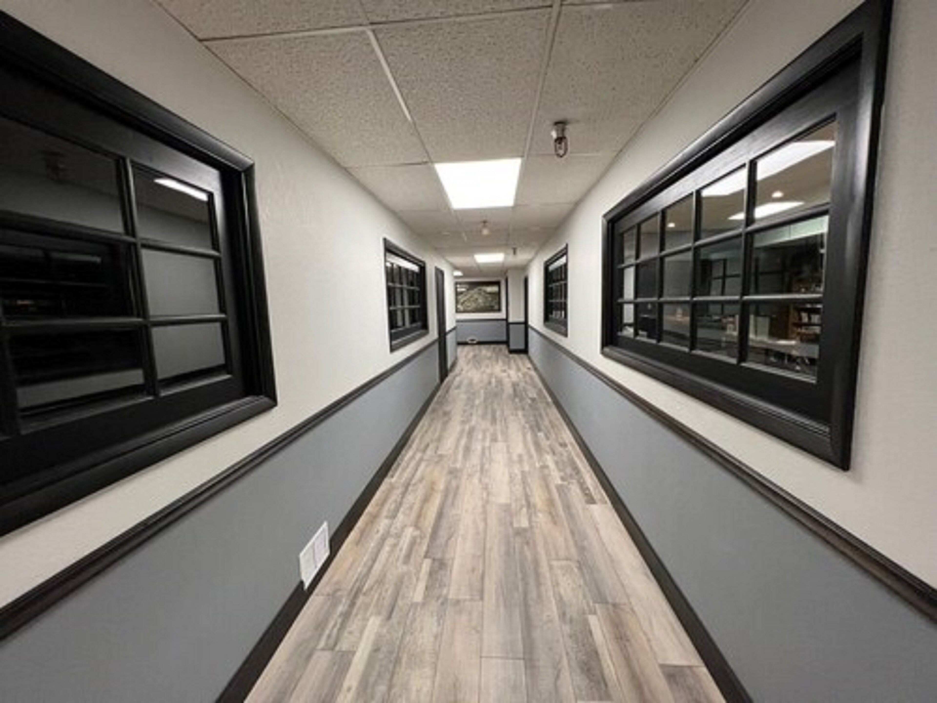 A long hallway with a wooden floor, large windows on both sides, and a ceiling with fluorescent lights.