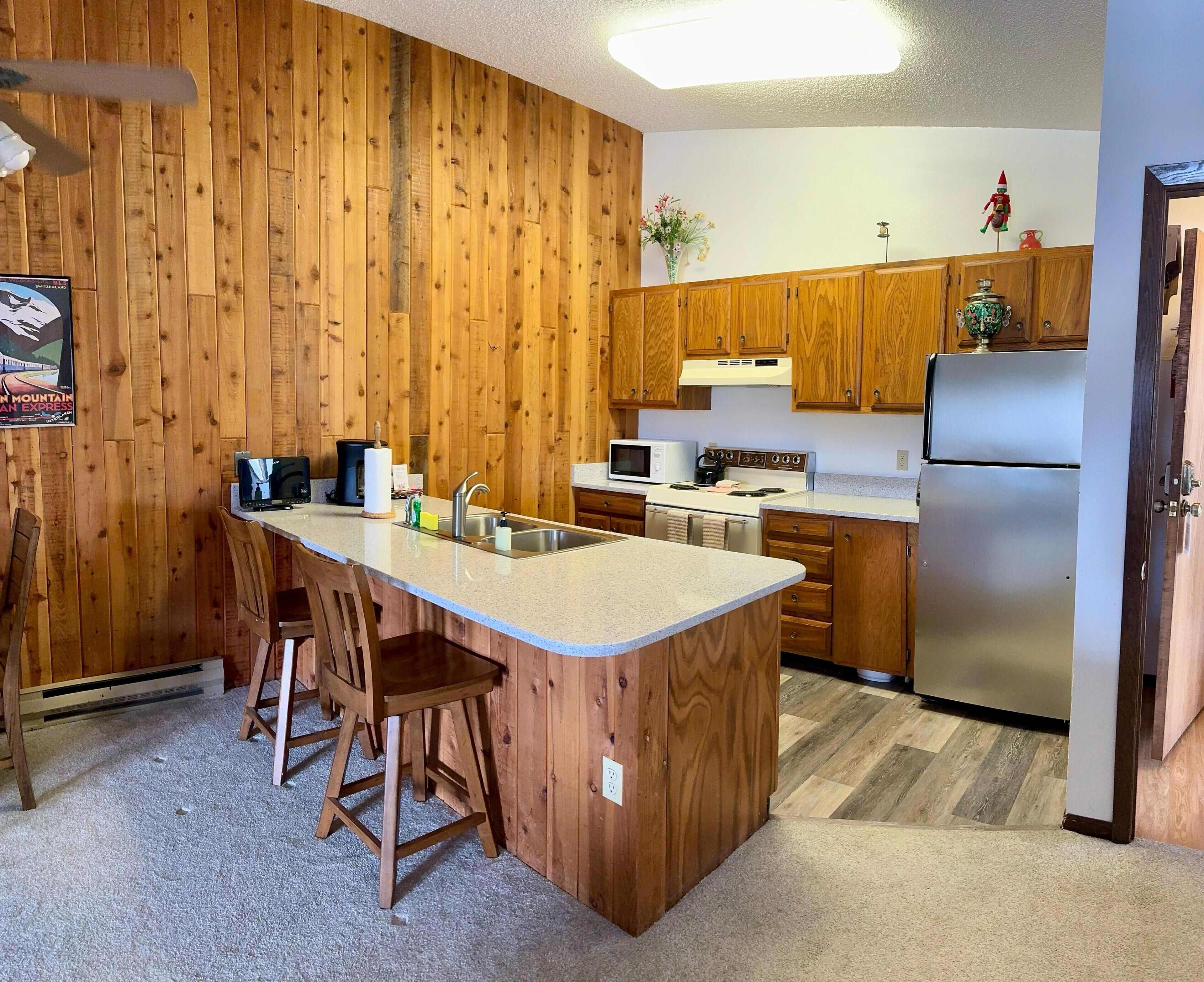 A kitchen with wooden panel walls, a central island with stools, a stainless steel refrigerator, and a microwave.