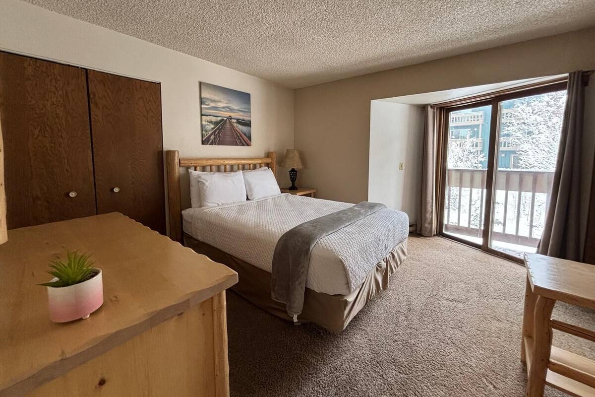 A hotel room with a large bed, a desk, a small plant, a painting on the wall, and a view through a large window.