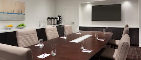 A conference room with a large table, chairs, a projector screen, a coffee station, and a painting on the wall.