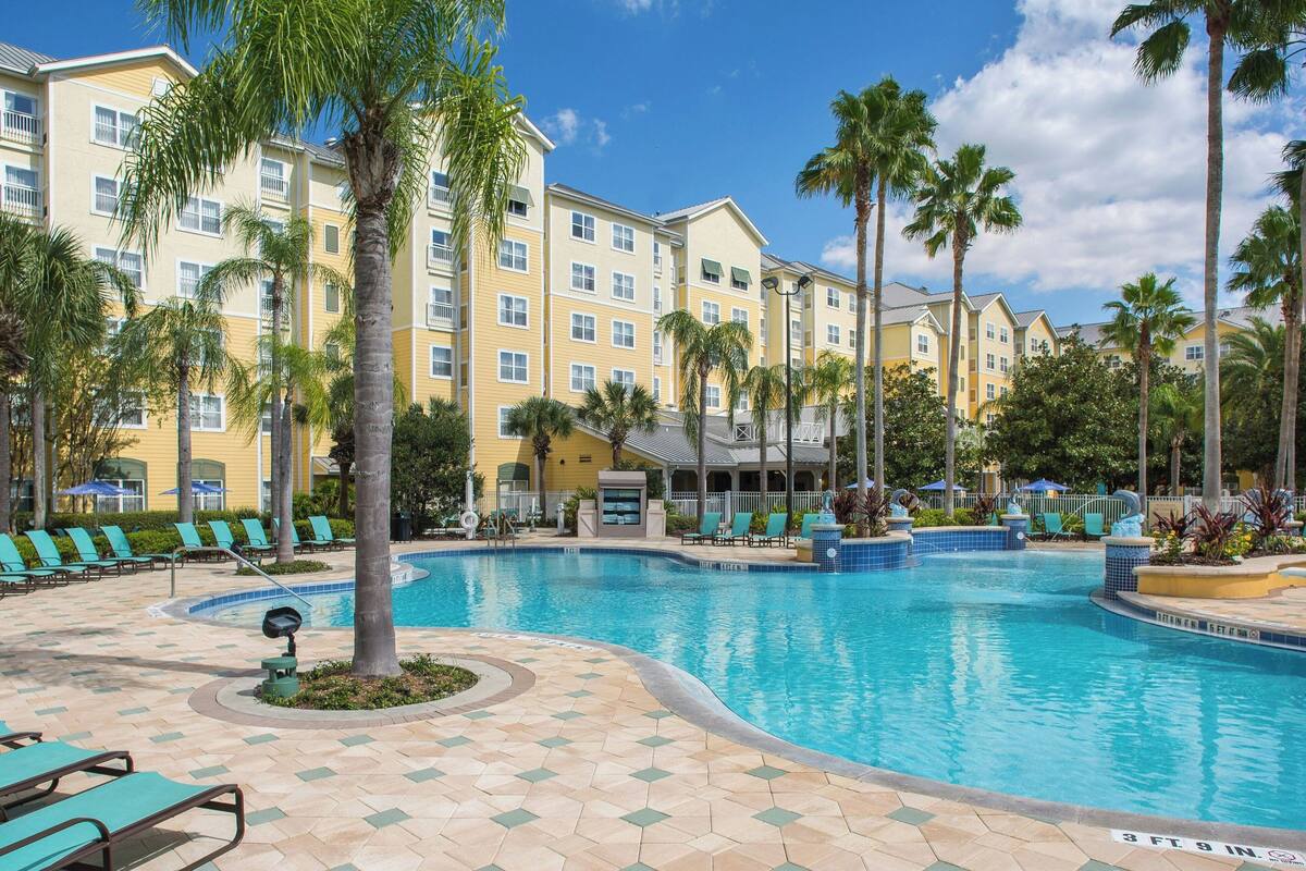 A swimming pool surrounded by palm trees and lounge chairs, with a hotel building in the background.