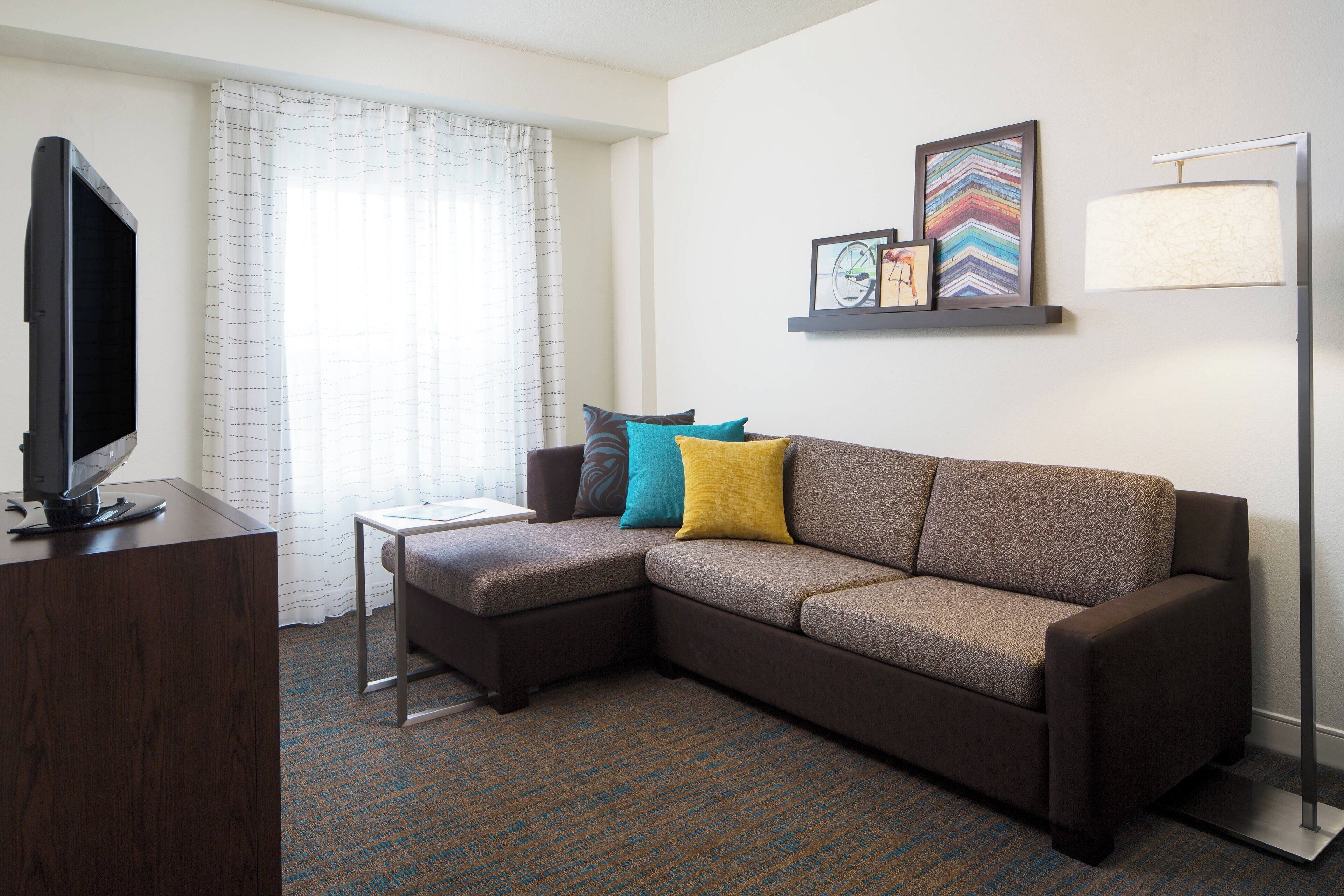 A modern hotel room with a sectional sofa, a flat-screen TV, a small table, and a window with curtains.