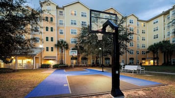 Quadra de basquete externa com cesta, cercada por um edifício com várias varandas e janelas.