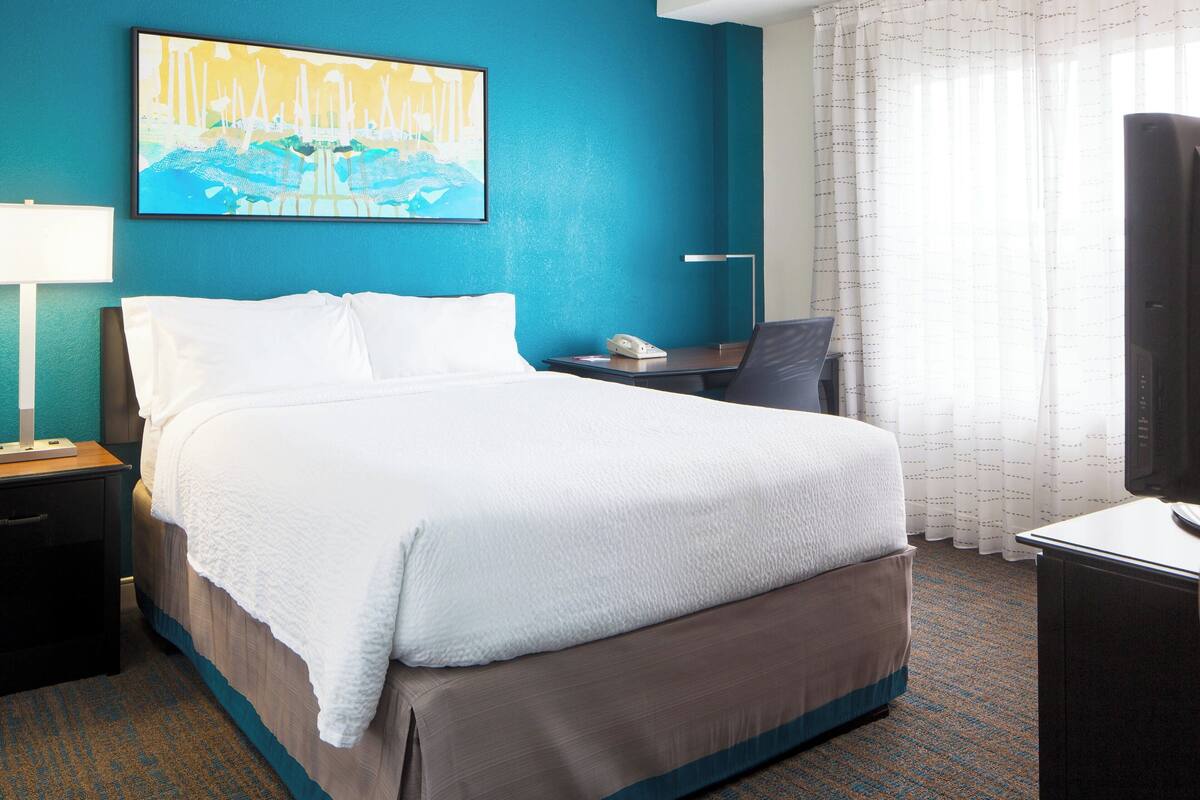 A hotel room with a large bed, a desk, a chair, a TV, and a painting on the wall.