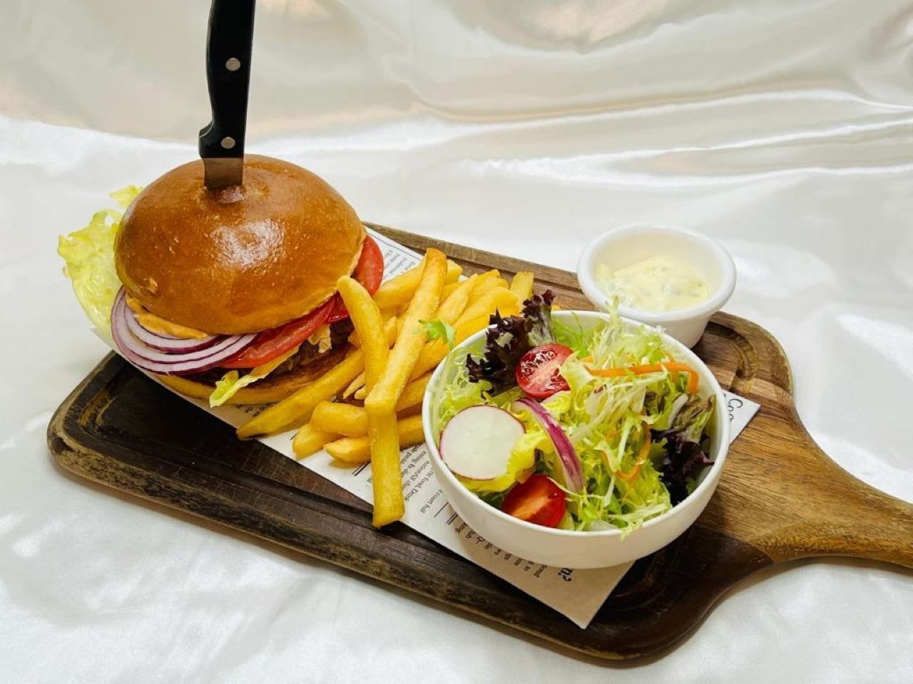 Un hamburger accompagné de frites et d’une salade, le tout servi sur une planche en bois.