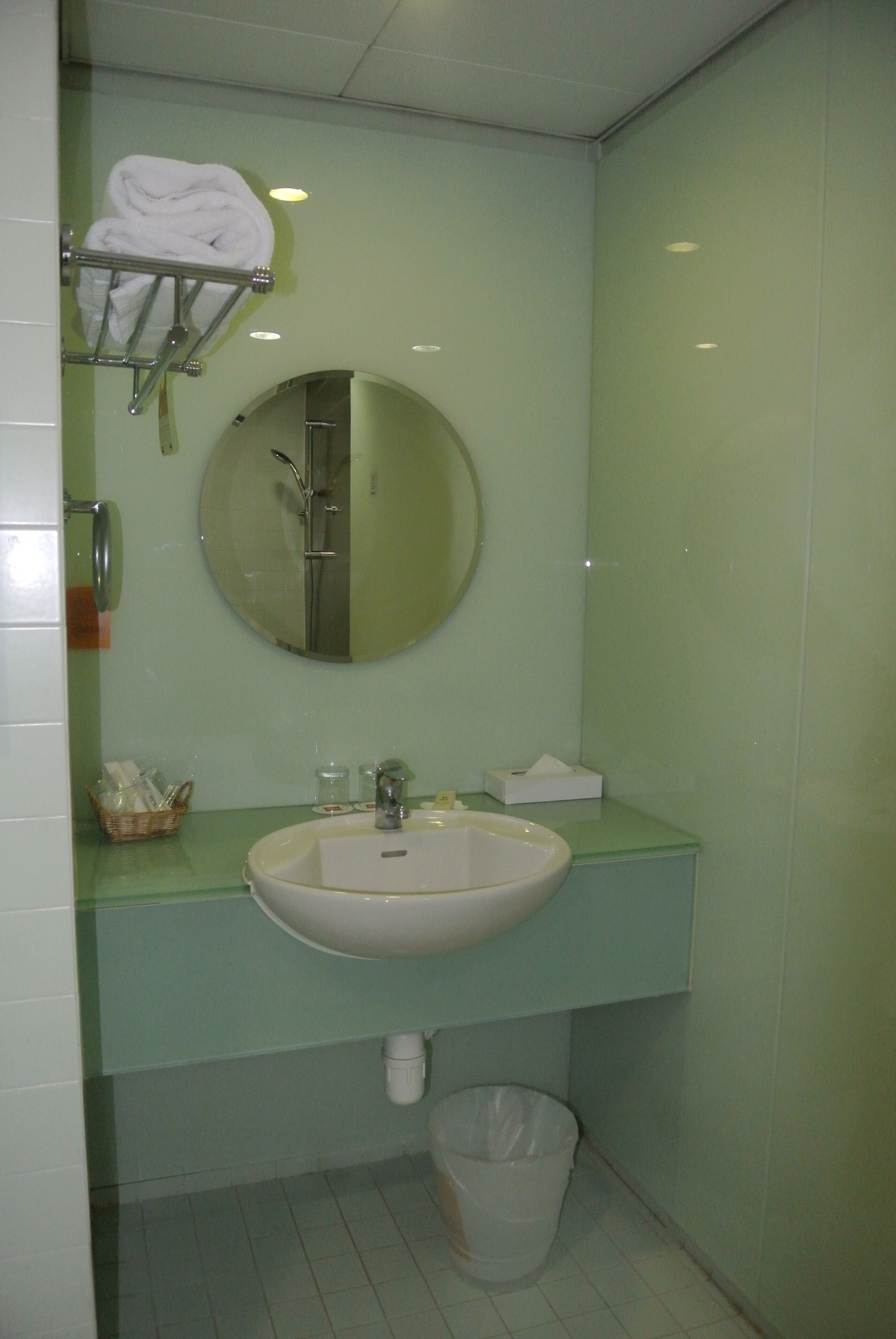Un baño con lavamanos, espejo, tendedero para toallas y un cubo de basura.