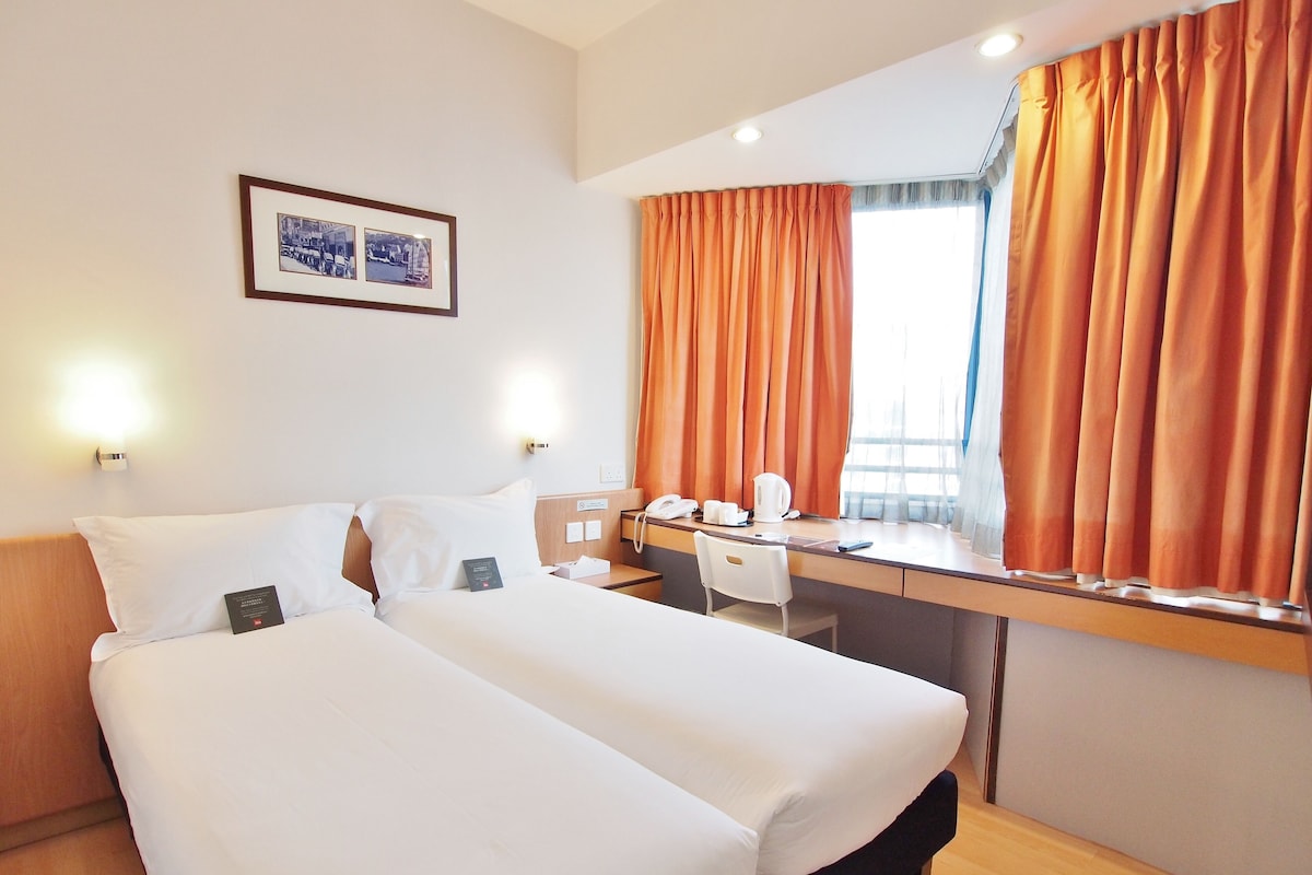 Habitación de hotel con dos camas, un escritorio con una silla, una ventana con cortinas naranjas y un cuadro en la pared.