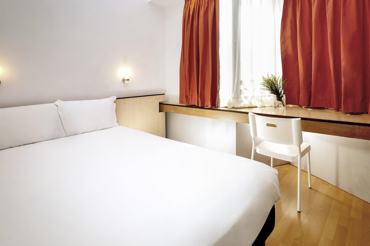 A hotel room with a bed, a desk, a chair, a window with curtains, and a wall-mounted light.
