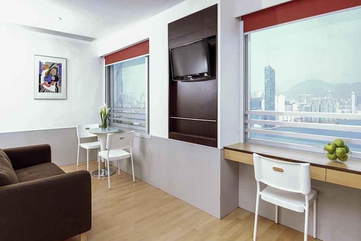 Una sala de estar moderna con un sofá, un escritorio con una silla, un televisor montado en la pared y un ventanal con vista a la ciudad.