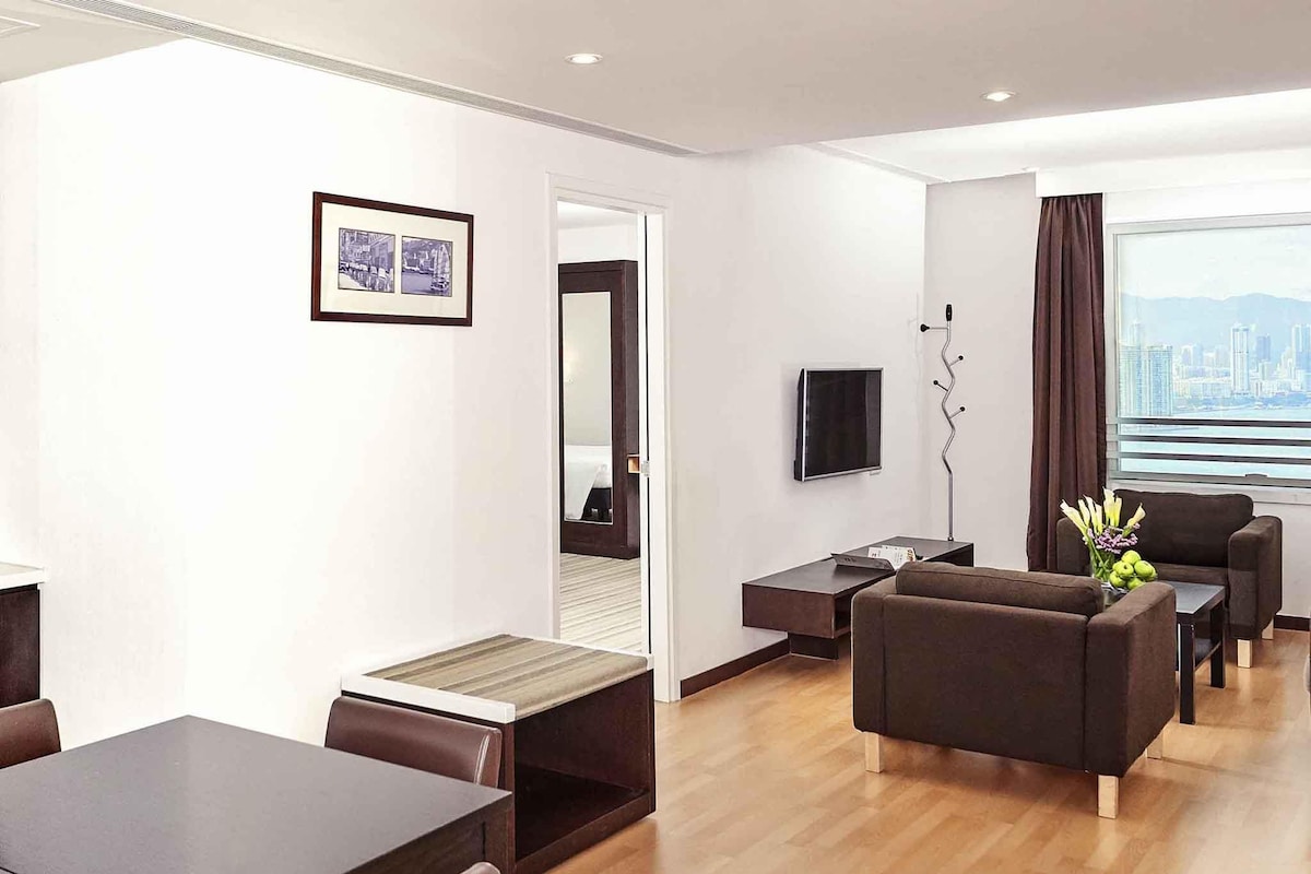 A modern hotel room with a dining area, a sofa set, a TV, and a city view.
