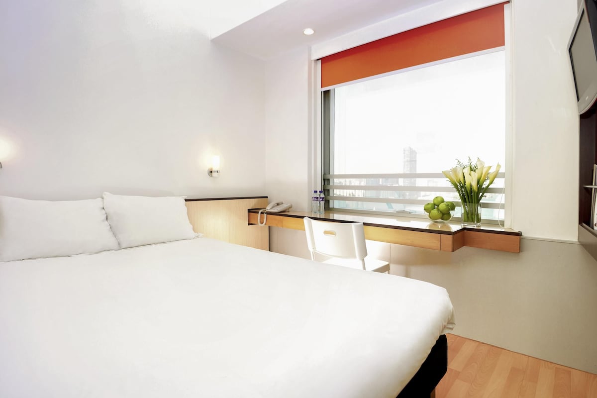 A hotel room with a large bed, a desk with a phone, a chair, a window with a view, and a vase with flowers.