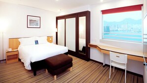 Una camera d'albergo con un letto, una scrivania, una sedia, un comodino, una lampada, un quadro incorniciato, un'ampia finestra vista città e un tappeto a righe.
