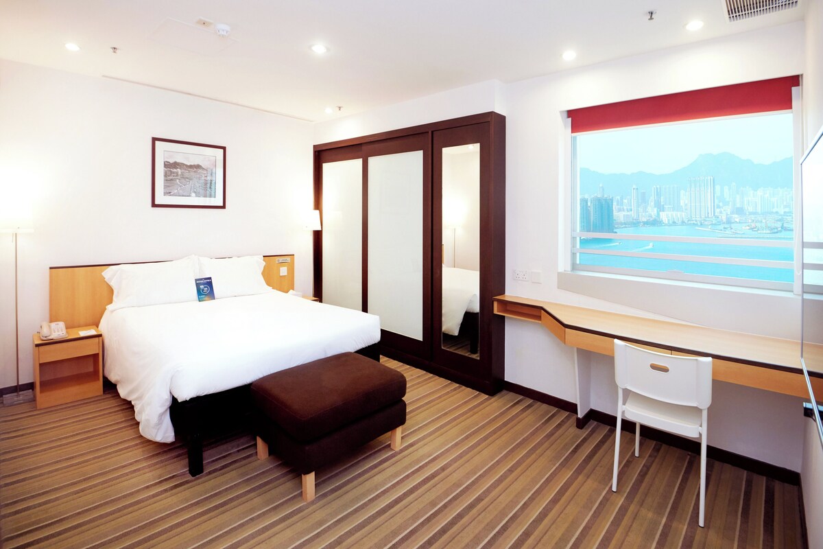 Habitación de hotel con cama, escritorio, silla, mesita de noche, lámpara, un cuadro enmarco, una ventana grande con vista a la ciudad y alfombra a rayas.