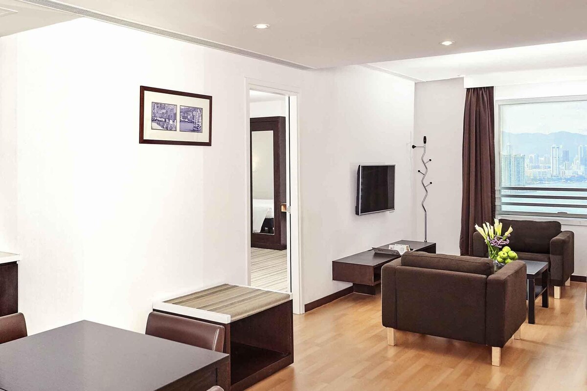 A modern hotel room with a dining area, a sofa set, a TV, and a city view.