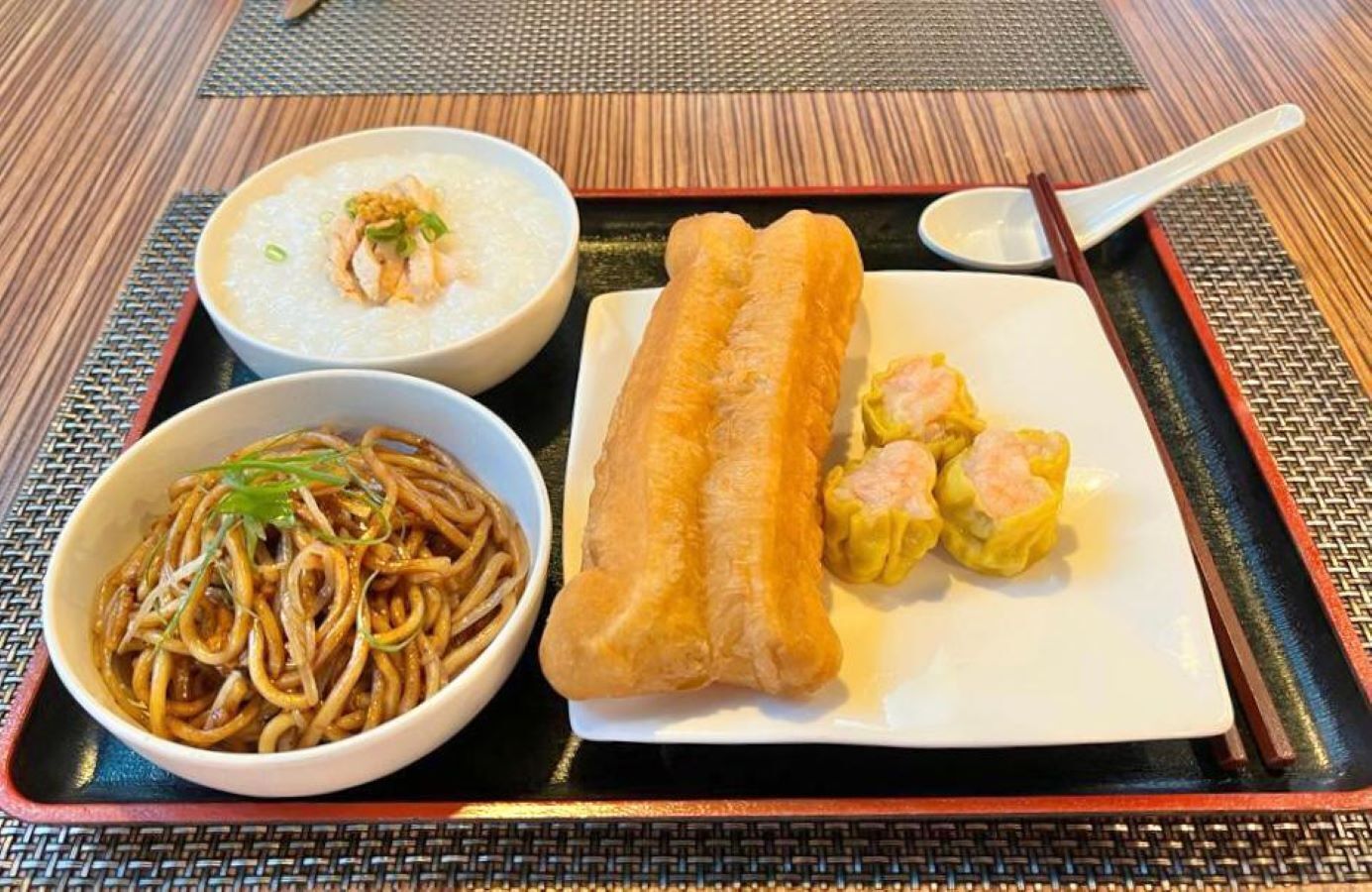 A meal consisting of a bowl of noodles, a bowl of soup, a piece of bread, and some dumplings on a tray.