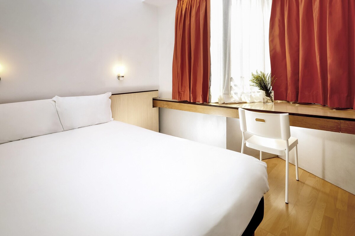 A hotel room with a bed, a desk, a chair, a window with curtains, and a wall-mounted light.
