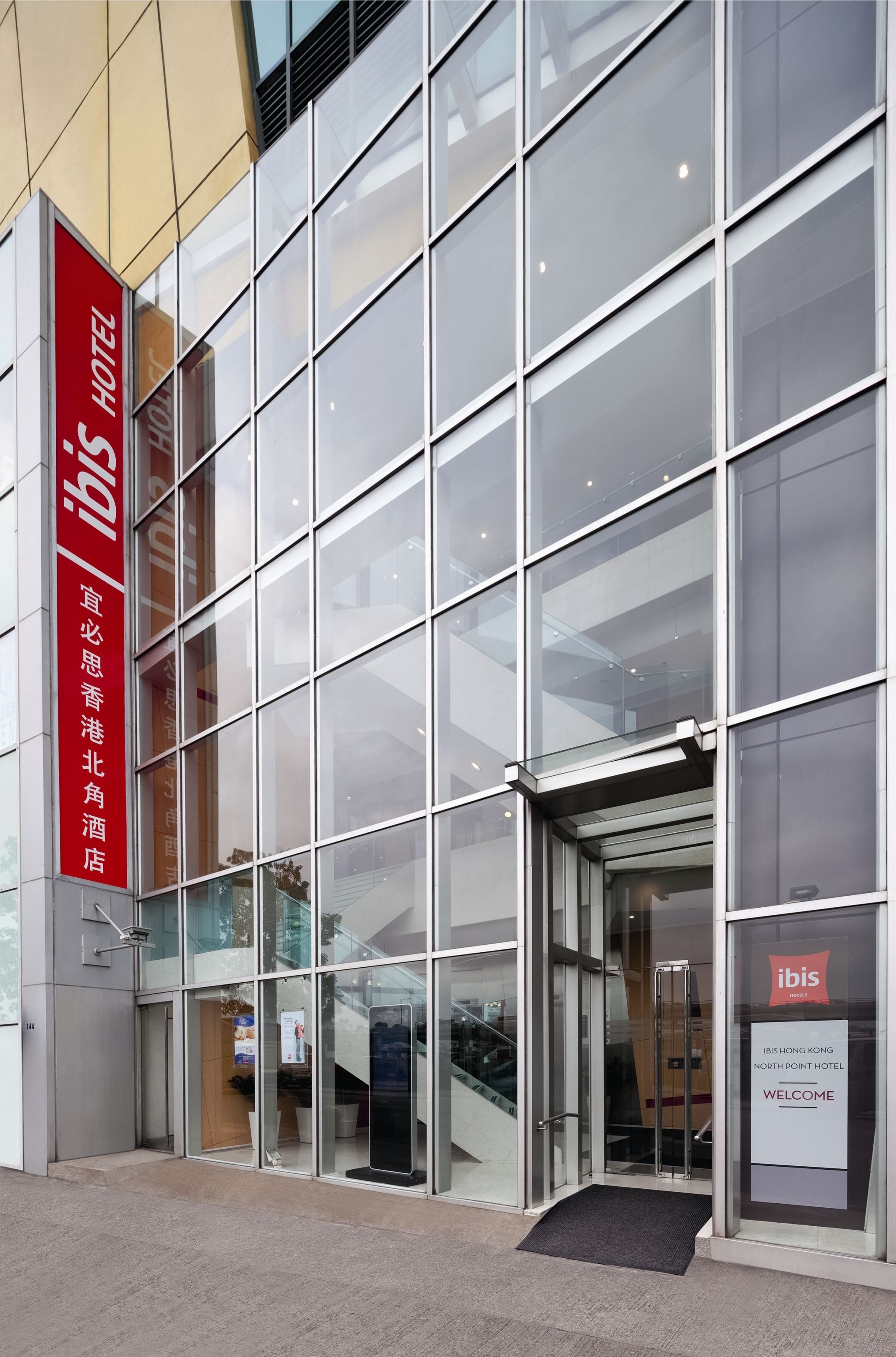 A modern hotel entrance with a glass facade, a red sign indicating the hotel name, and a welcome sign.