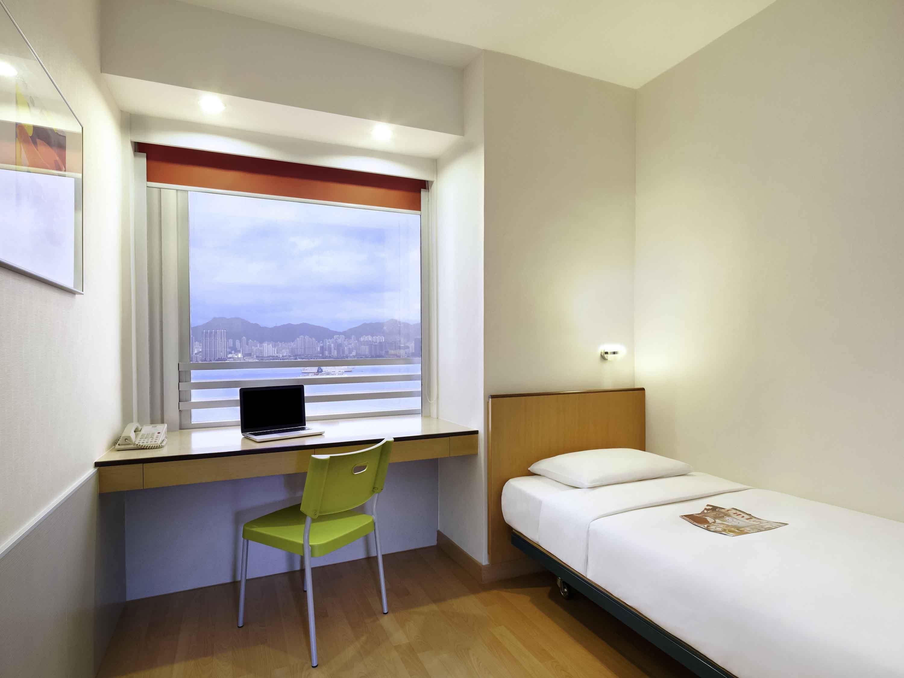 Een hotelkamer met een bed, een bureau met een laptop, een stoel, een raam met uitzicht over de stad en een telefoon op het bureau.
