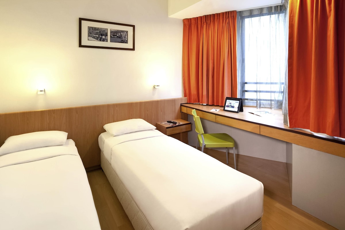 A hotel room with two beds, a desk with a chair, a window with orange curtains, and a framed picture on the wall.