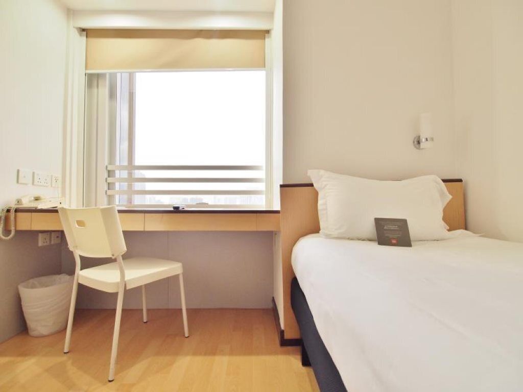 A hotel room with a bed, a desk with a chair, a window with blinds, and a wall-mounted light.