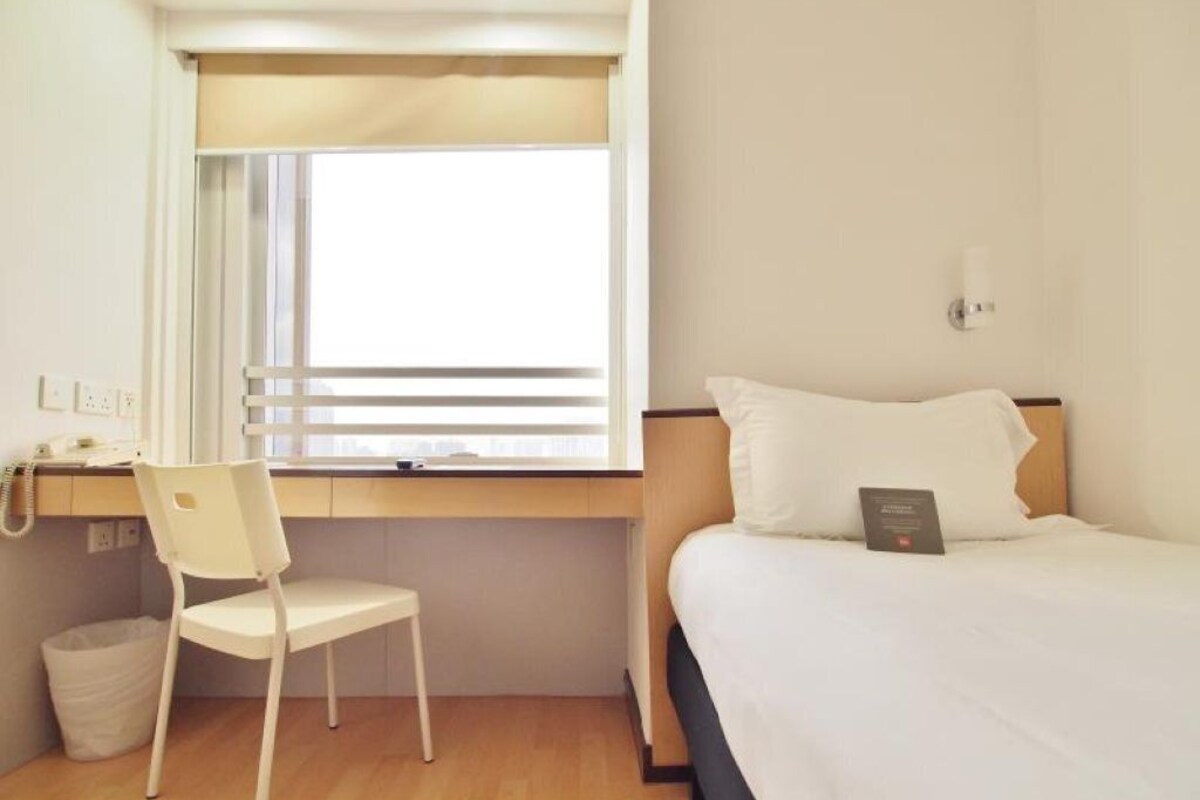 Habitación de hotel con cama, escritorio, silla, ventana con persianas y una lámpara de pared.