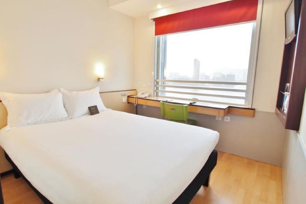 Habitación de hotel con una cama grande, un escritorio, una silla, una ventana con persianas y vistas a la ciudad.