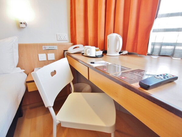 A hotel room with a wooden desk, a white chair, a telephone, a coffee maker, a remote control, and a window with orange curtains.