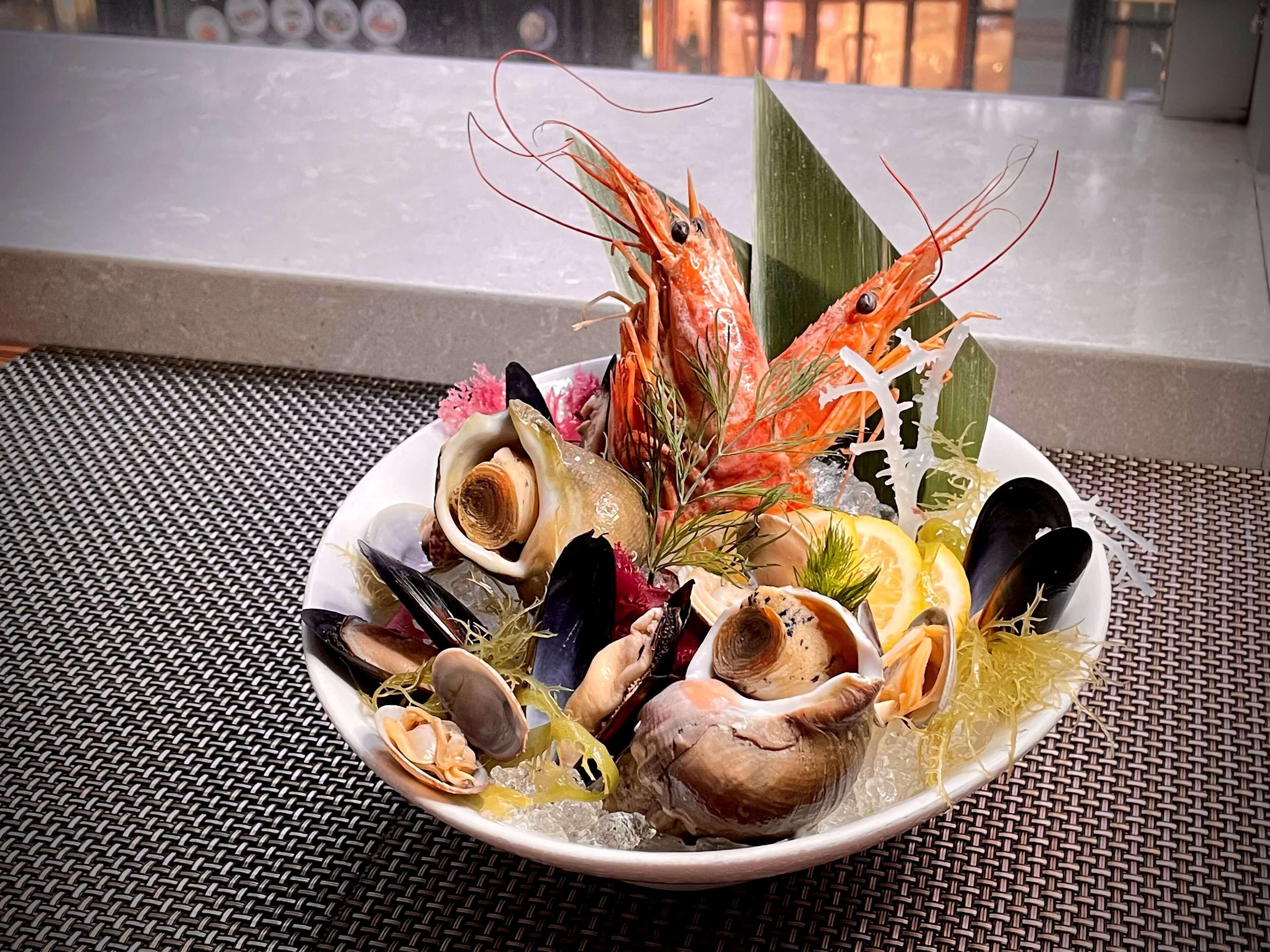 Una bandeja de mariscos con diversos crustáceos y un camarón decorativo sobre una estera trenzada.