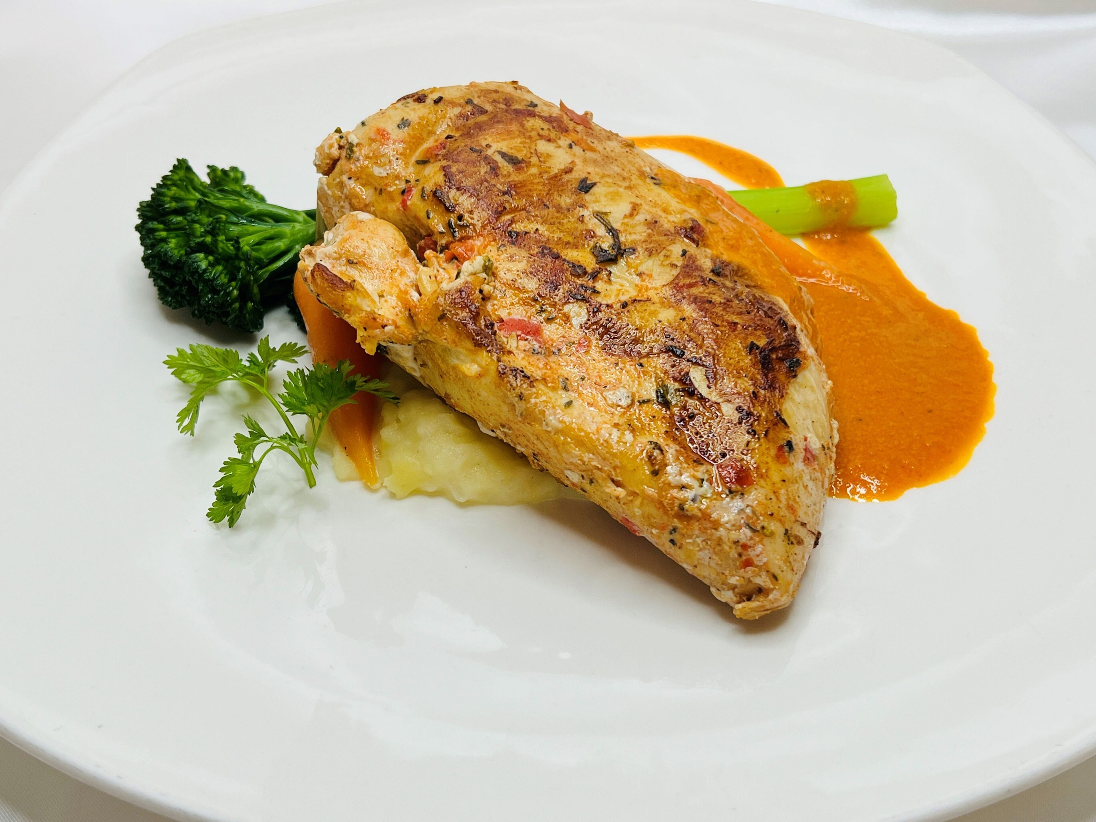 A plate with a piece of grilled chicken, mashed potatoes, broccoli, and a green vegetable, possibly asparagus, with a side of orange sauce.
