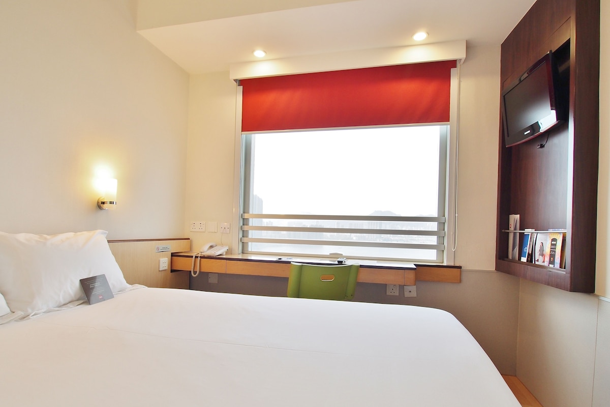 Habitación de hotel con cama, escritorio con teléfono, ventana con persianas y televisor montado.