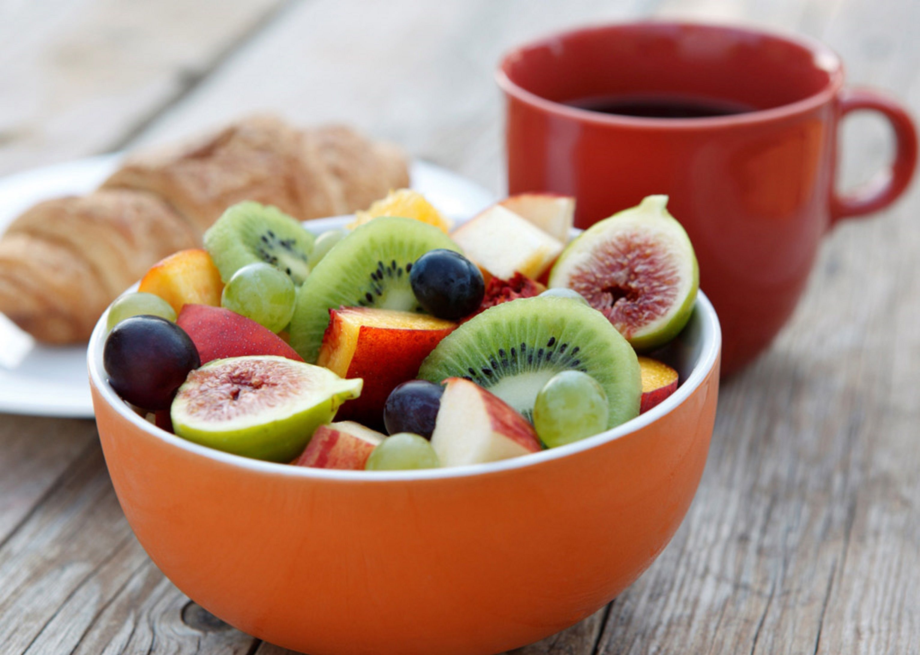 Uma tigela com frutas variadas e uma caneca vermelha sobre uma mesa de madeira.