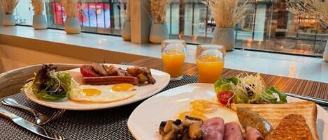 A breakfast setting with plates of eggs, bacon, mushrooms, ham, and toast, along with a side salad and orange juice.