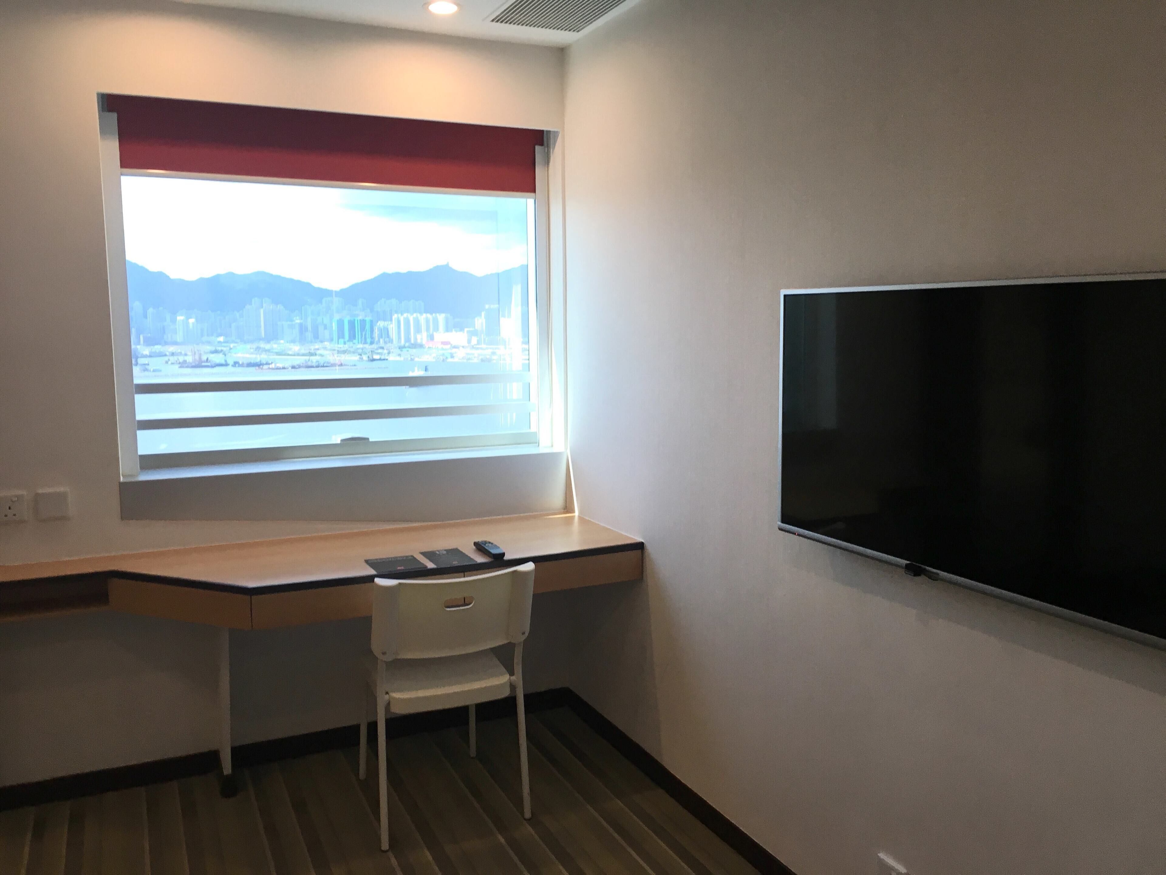 A room with a desk, a chair, a TV, and a window with a view of a cityscape.