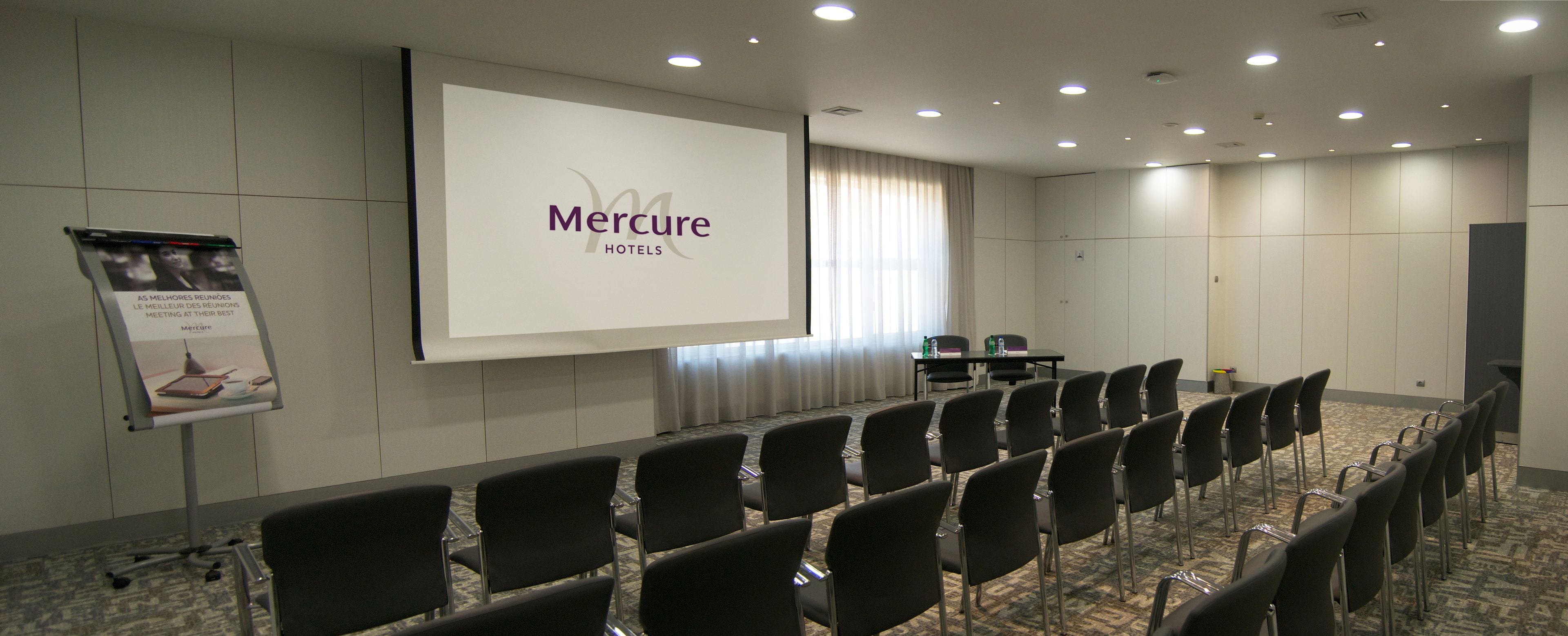 Ein Konferenzraum mit einer großen Leinwand, die die Marke 'Mercure Hotels' zeigt, Reihen schwarzer Stühle, einem kleinen Tisch mit einer Flasche und einem Teppichboden.