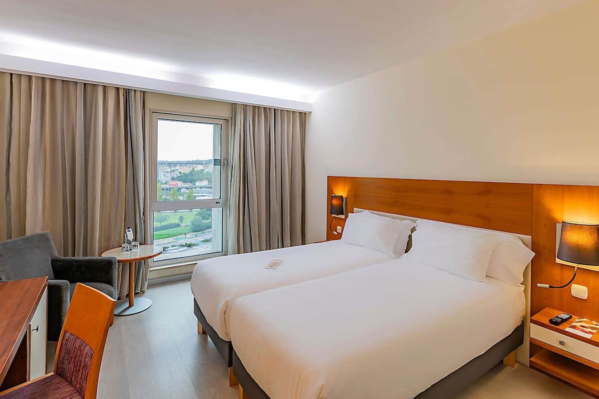 Ein Hotelzimmer mit einem großen Bett, einem Schreibtisch, einem Sessel, einem kleinen Tisch, einer Lampe und Blick auf die Stadt durch das Fenster.