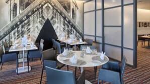 A dining area with round tables set for four, each with white tablecloths, napkins, and cutlery. The room features a large, textured wall mural and a patterned carpet.