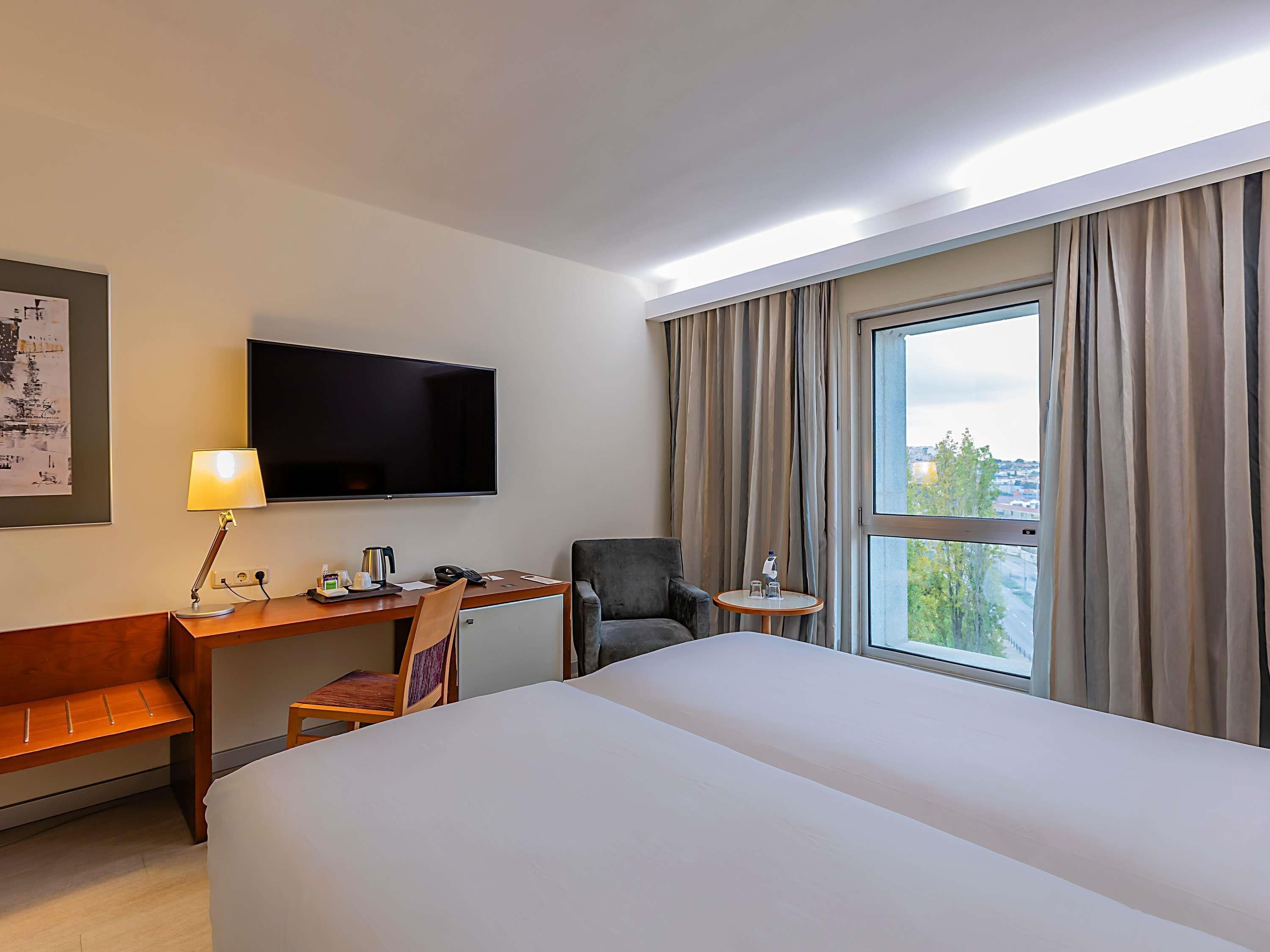A hotel room with a bed, desk, chair, TV, and a window with curtains.