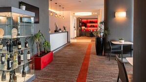 A hotel lobby with a reception desk, glass display cases with bottles, a red planter with plants, a carpeted floor, and a dining area with chairs and a table. - Mercure Lisboa Hotel (Lisbon)