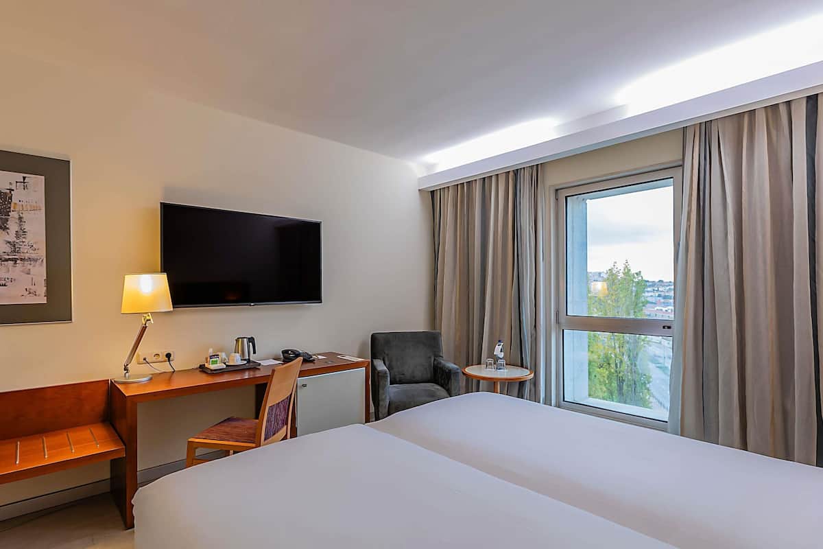Habitación de hotel con cama, escritorio, silla, televisor y ventana con cortinas.