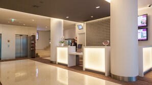 A modern hotel lobby with a reception desk, a digital screen, and an elevator.