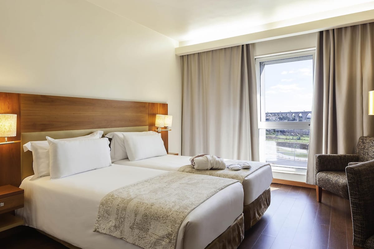 Habitación de hotel con dos camas, un ventanal con vistas, una silla y una mesita de noche con lámpara.
