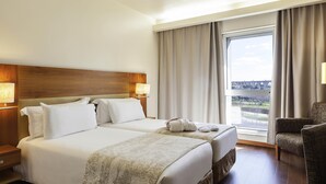 A hotel room with two beds, a large window with a view, a chair, and a nightstand with a lamp. - Mercure Lisboa Hotel (Lisbon)