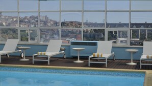 A rooftop pool area with lounge chairs and a view of the cityscape through large windows.