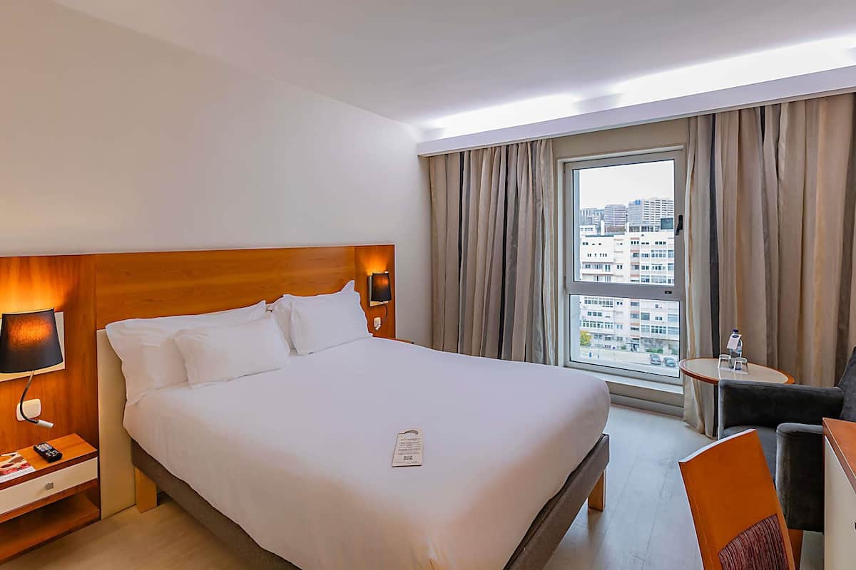 Habitación de hotel con una cama grande, una mesita de noche con lámpara, una pequeña mesa con una silla y una ventana con cortinas.