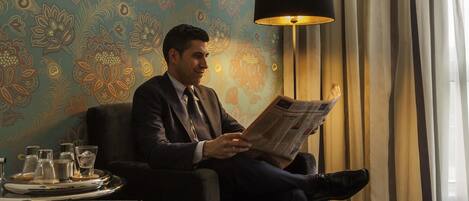 A man seated in a room with a patterned wallpaper, a lamp, and a tray with glasses and a bottle.