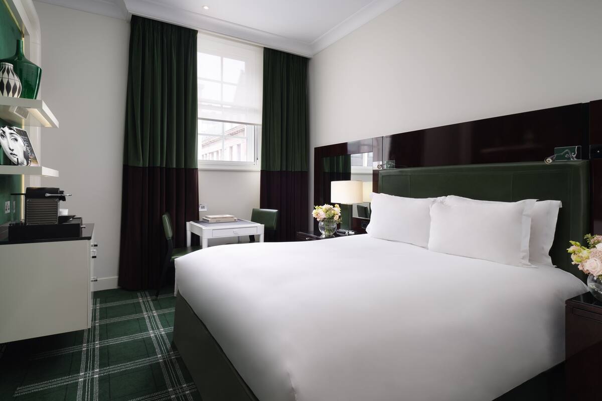A hotel room with a large bed, a desk, a chair, a nightstand with a lamp, and a window with curtains.