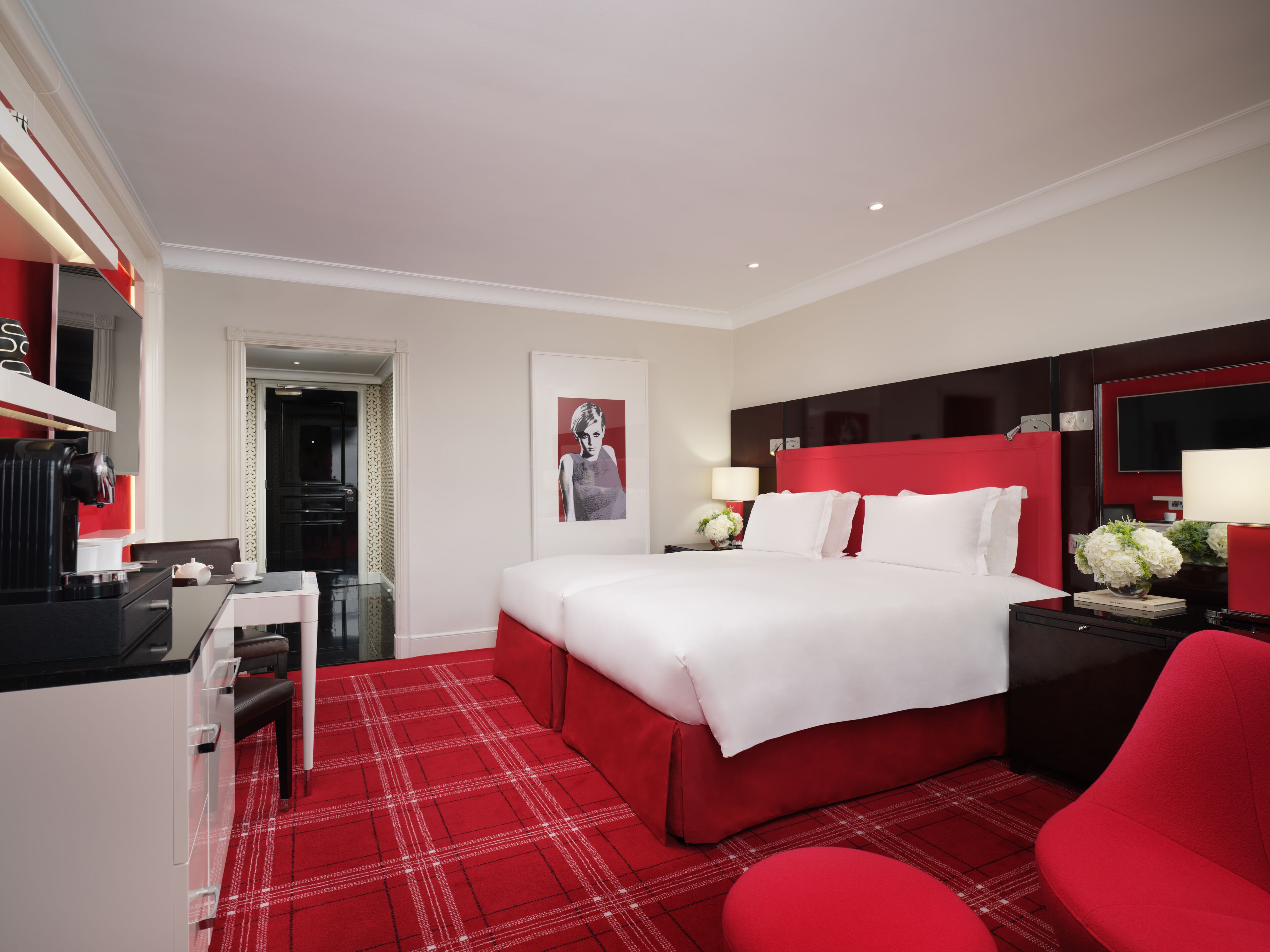 A hotel room with a red and white color scheme, a bed with white linens, a red headboard, a bedside table with a lamp, and a red armchair.