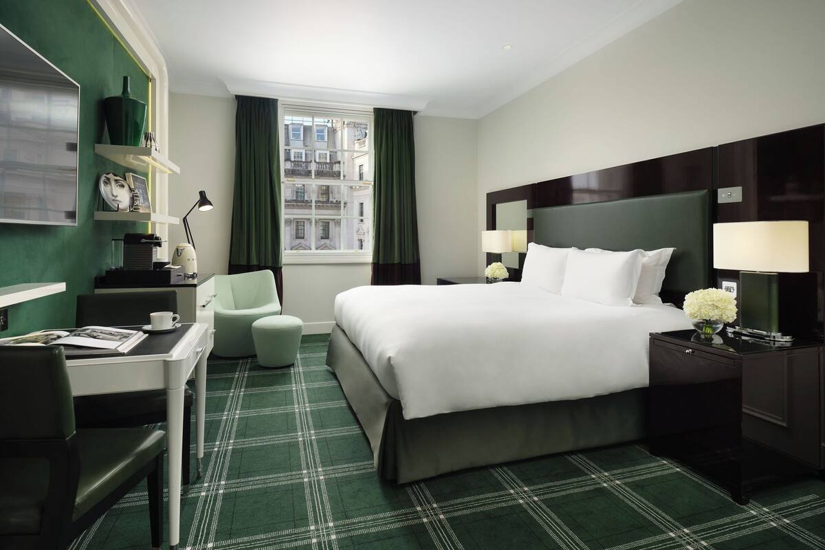 A hotel room with a bed, a desk, a chair, a nightstand, a lamp, and a window with curtains.