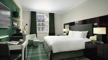 Quarto de hotel com cama, escrivaninha, cadeira, mesa de cabeceira, luminária e janela com cortinas.